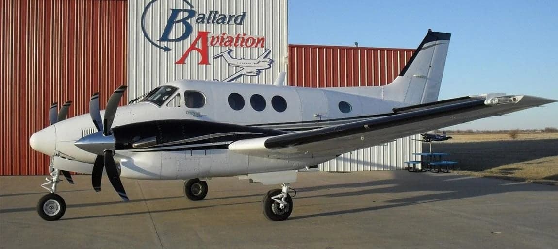 A Beechcraft King Air is parked in front of a hangar with the Ballard Aviation logo.