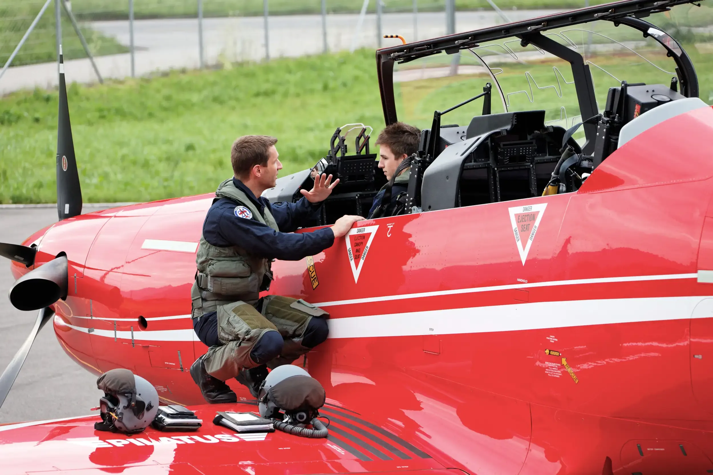 A pilot talking to a person in the cockpit of a red Pilatus PC-21 aircraft on the ground.
