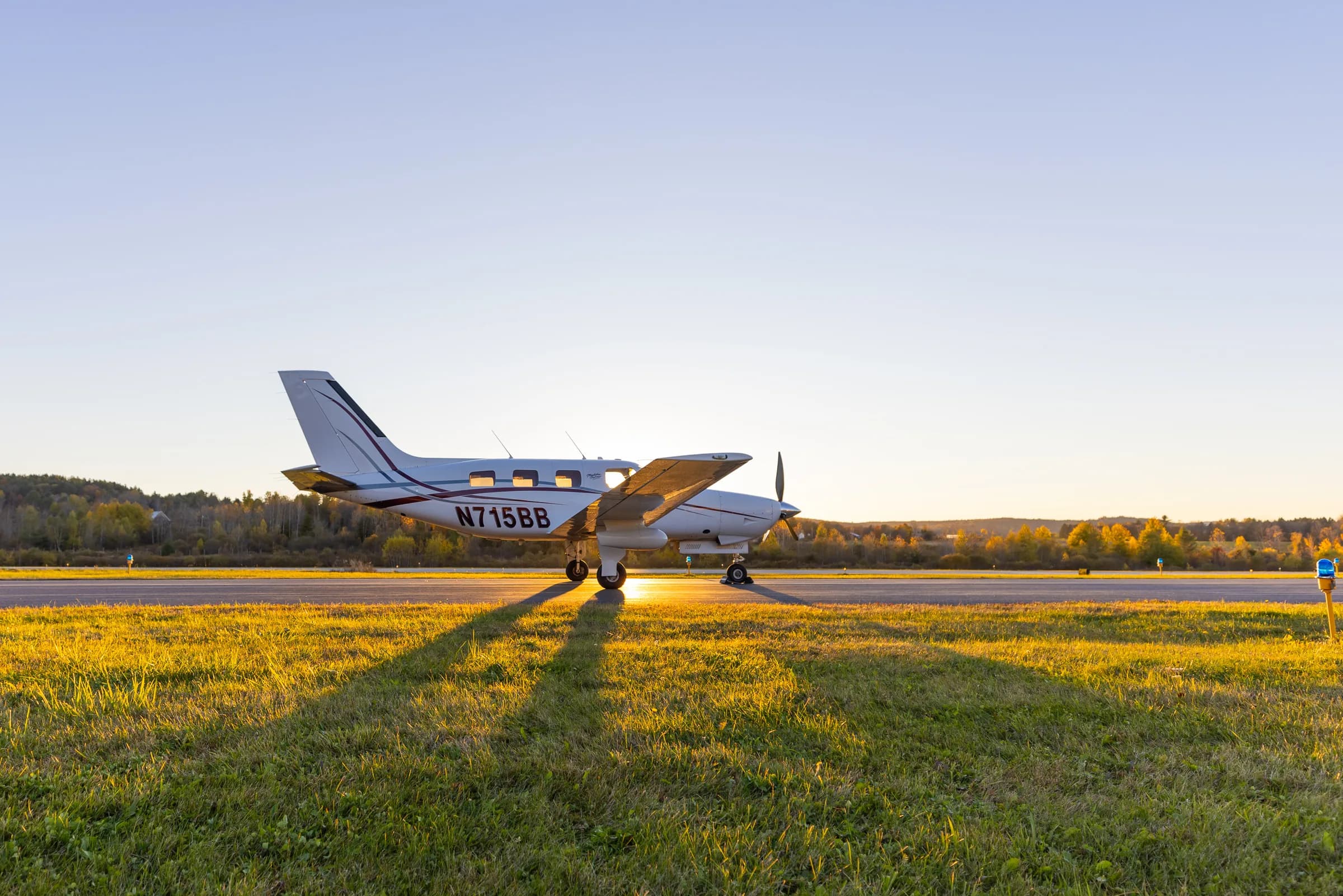 A white Piper Malibu Mirage N715BB with red and grey stripes on a runway at sunset.