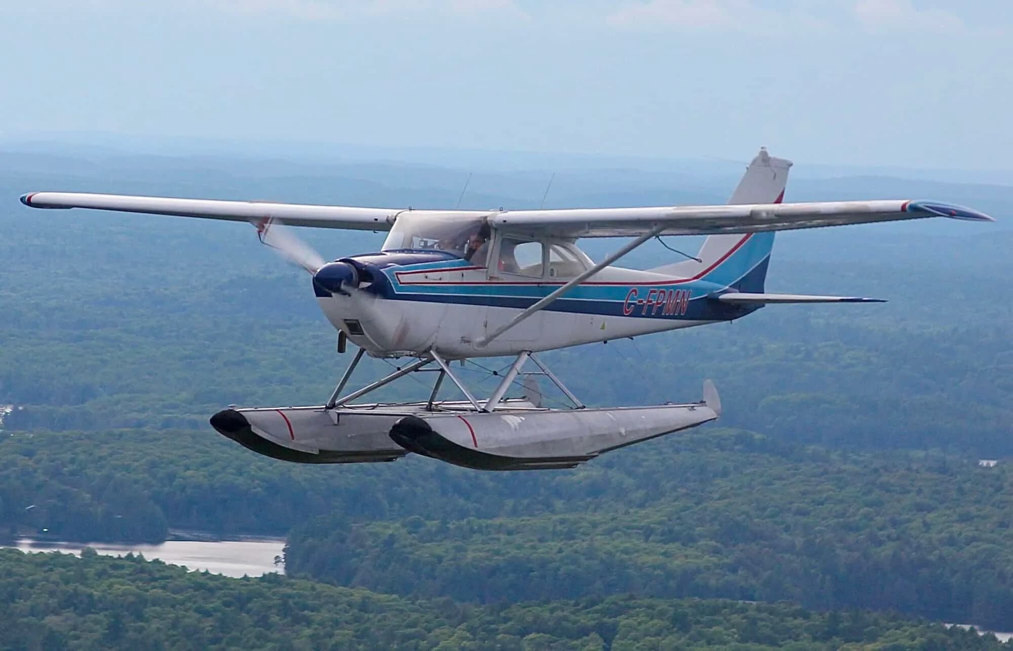 A white and blue Cessna 172 floatplane, registration C-FPMN, flies over a forested landscape with water below.