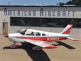A white and red single-engine Piper aircraft, registration N39954, parked on a tarmac in front of an American Aircraft hangar.
