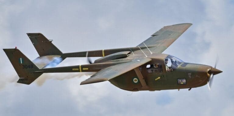 A military-style aircraft with tail number 3048 in flight against a cloudy sky.
