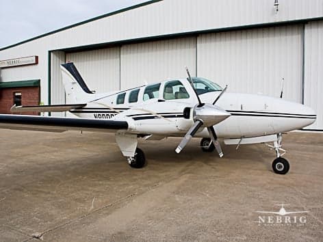 A white twin-engine propeller aircraft with black stripes is parked on a concrete apron in front of a hangar.
