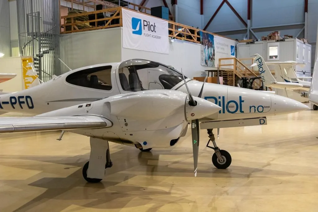 A white Diamond DA42 aircraft with blue "Pilot.no" livery parked in a hangar.