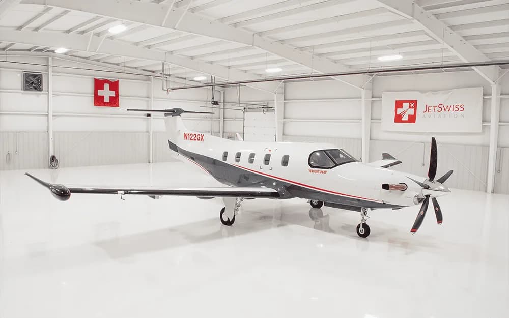 A white and grey Pilatus PC-12 turboprop aircraft with tail number N122GX parked inside a bright white hangar.