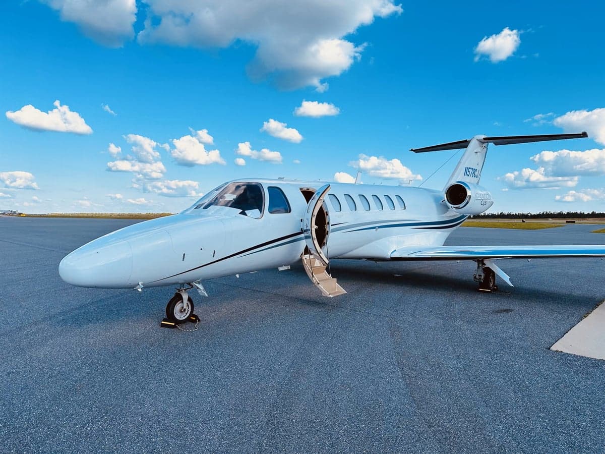 A white Cessna Citation CJ3 (N578CJ) with blue and grey stripes on the tail and fuselage, parked on an asphalt tarmac with its main door open, under a blue sky with white clouds.