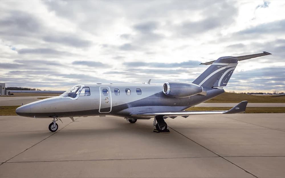 A silver and dark grey business jet is parked on an asphalt tarmac under a cloudy sky.