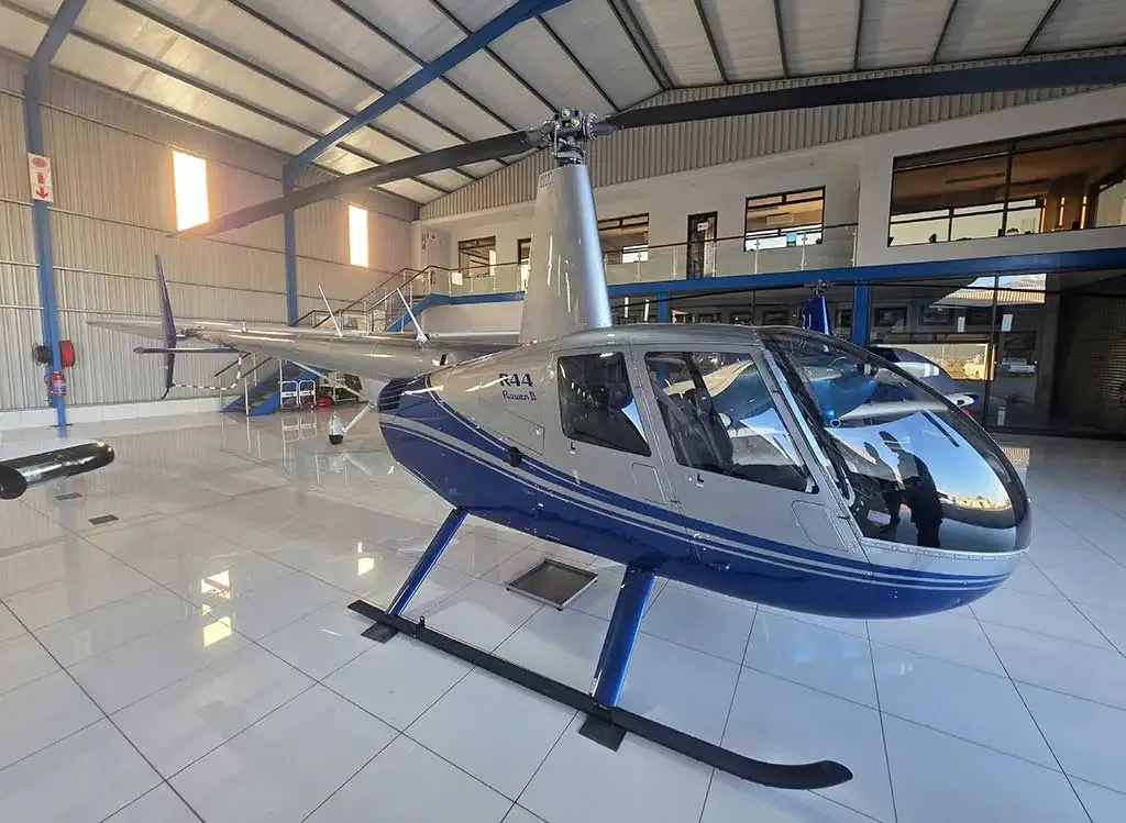 A silver and blue Robinson R44 Raven II helicopter parked inside a well-lit hangar.