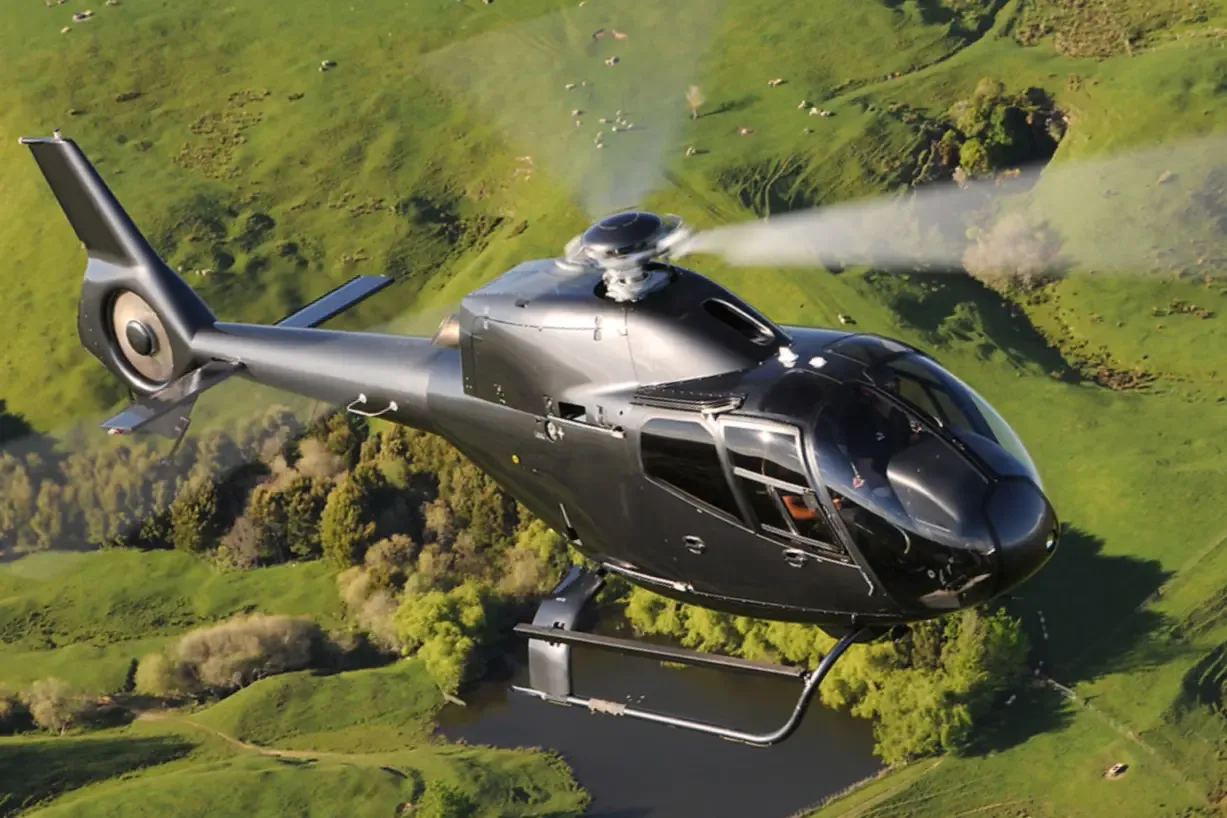 A sleek black helicopter with a fenestron tail rotor flies over lush green hills and a body of water.