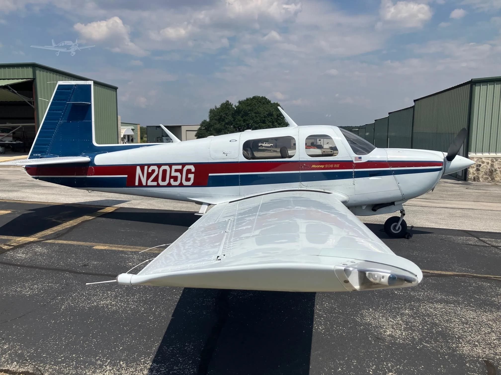 A white Mooney 205 SE aircraft with red and blue stripes is parked on an asphalt ramp with hangars in the background.