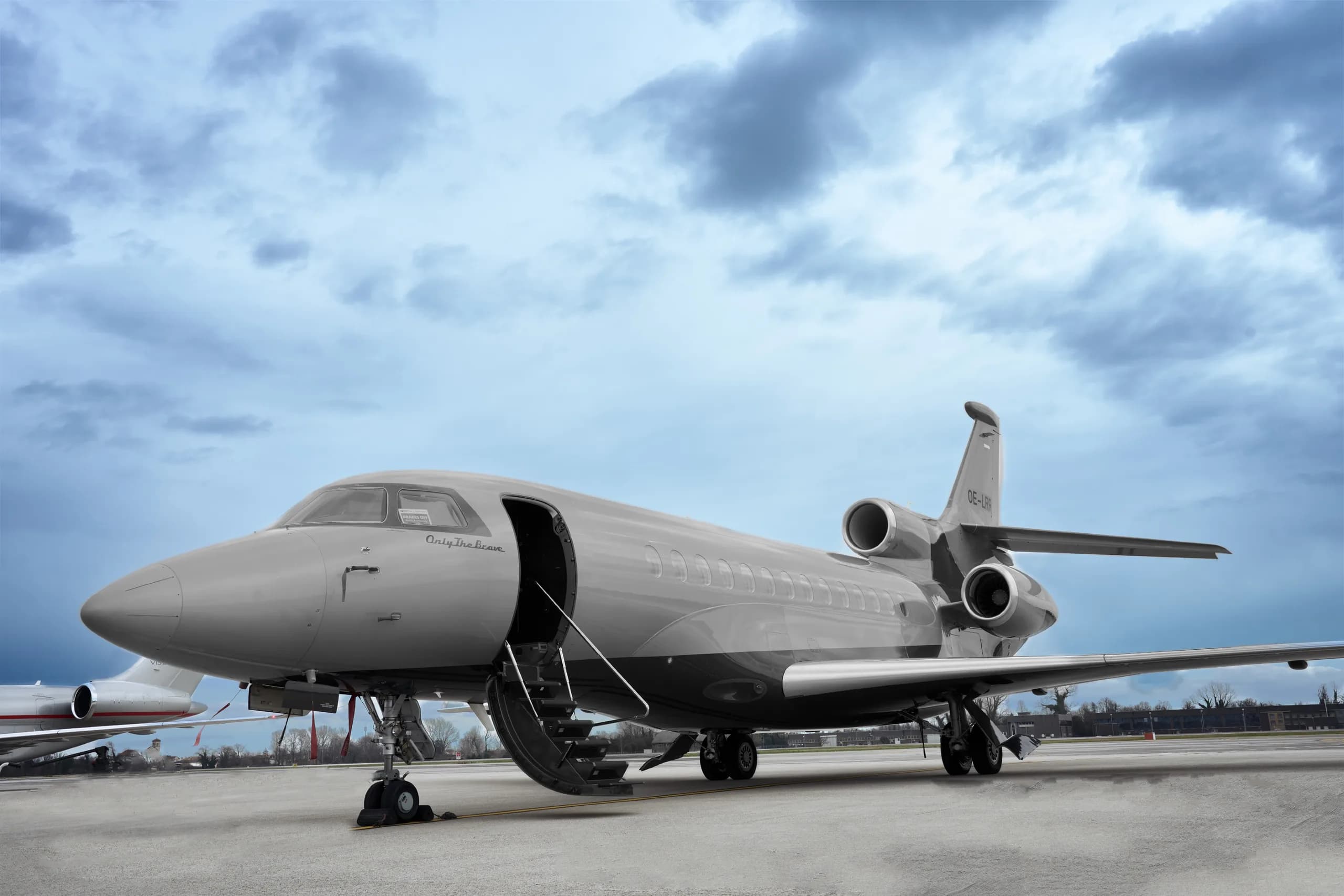 A grey Dassault Falcon 900EX business jet with its door open and stairs extended, parked on an airfield under a cloudy sky.