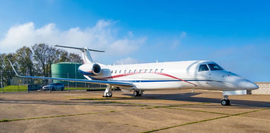 A white Embraer Legacy business jet with red and blue stripes parked on an airfield under a clear blue sky.