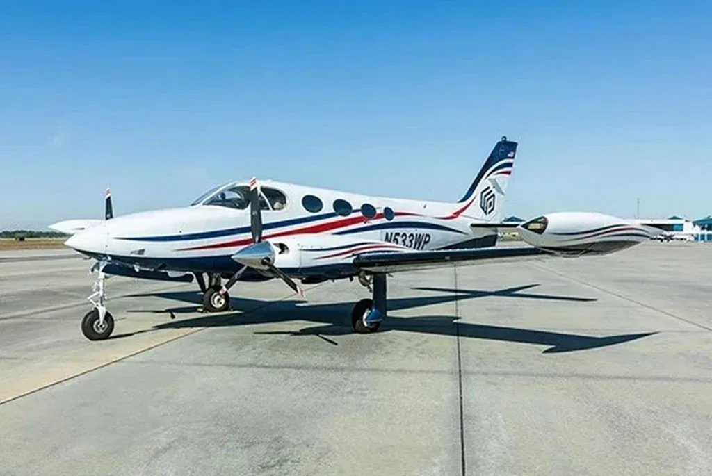A white twin-engine propeller aircraft with blue and red stripes on the fuselage and tail, parked on an airfield.