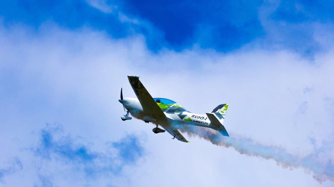 A white and green aircraft flies through the sky, leaving a smoke trail.