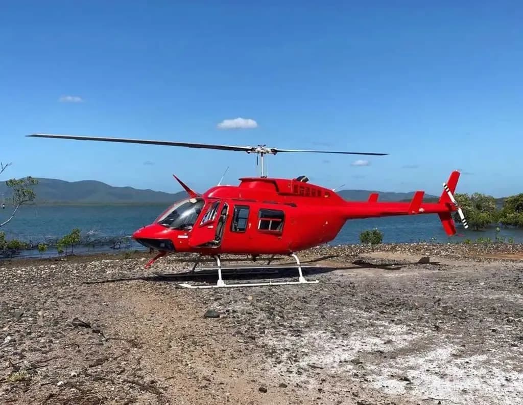 A bright red helicopter with white skids is parked on a dirt bank next to a body of water with trees and hills in the background.