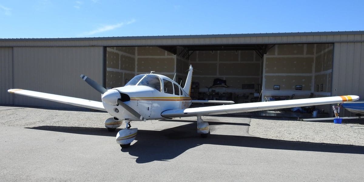 Front view of a white and yellow Piper Archer II aircraft parked in front of an open hangar.