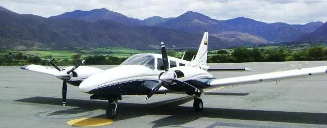A white and blue twin-engine propeller aircraft parked on an tarmac with mountains in the background.