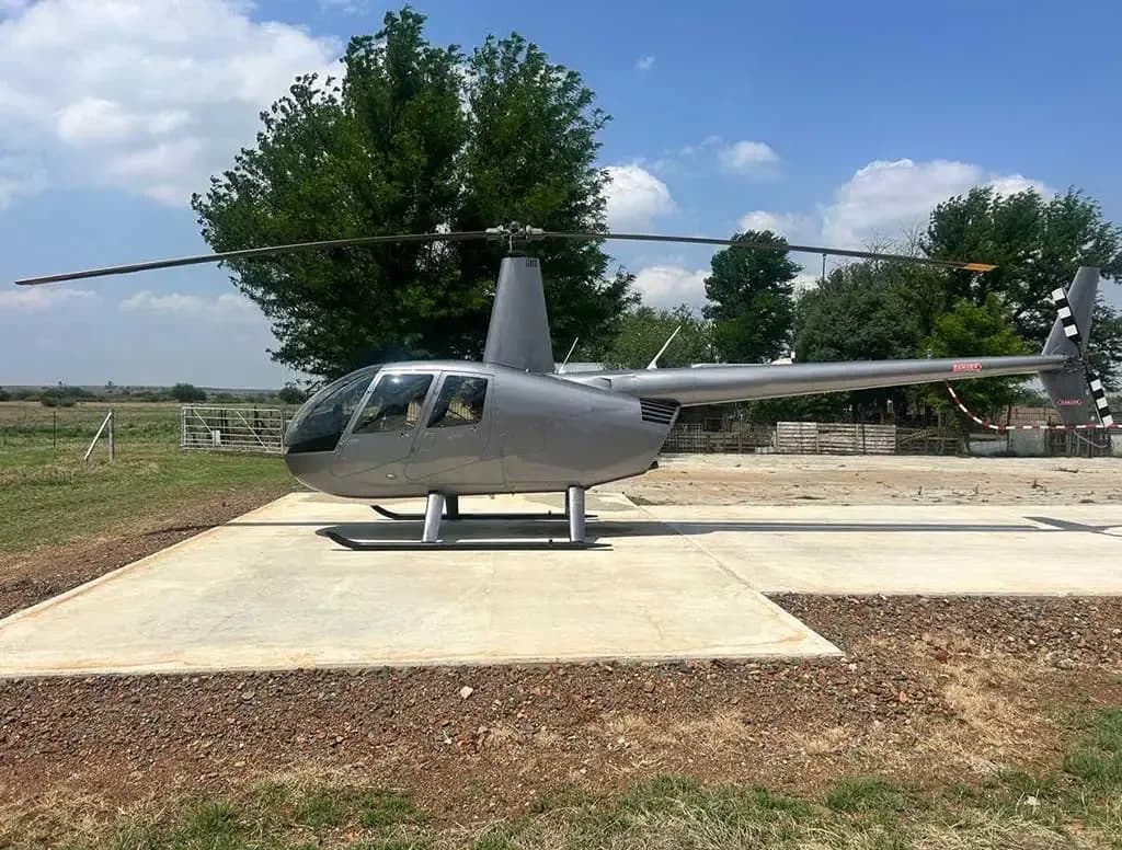 A silver Robinson R44 helicopter parked on a concrete pad in a grassy field under a partly cloudy sky.