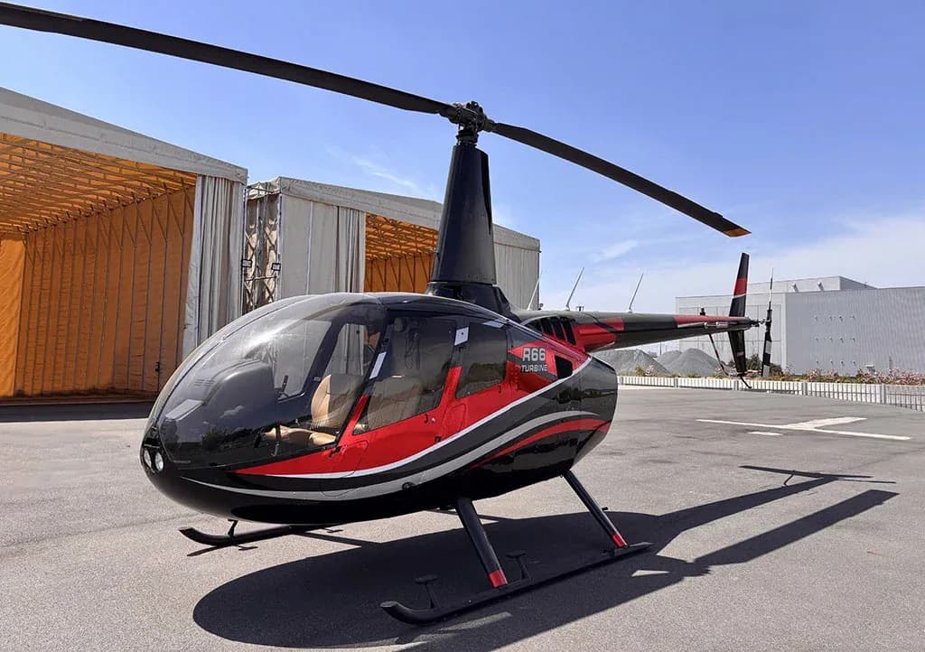 A black and red Robinson R66 Turbine helicopter with a silver stripe sits on an asphalt pad in front of hangars.