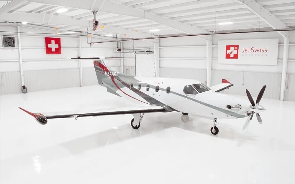 A white Pilatus PC-12 NGX aircraft with red and grey accents is parked in a brightly lit hangar.