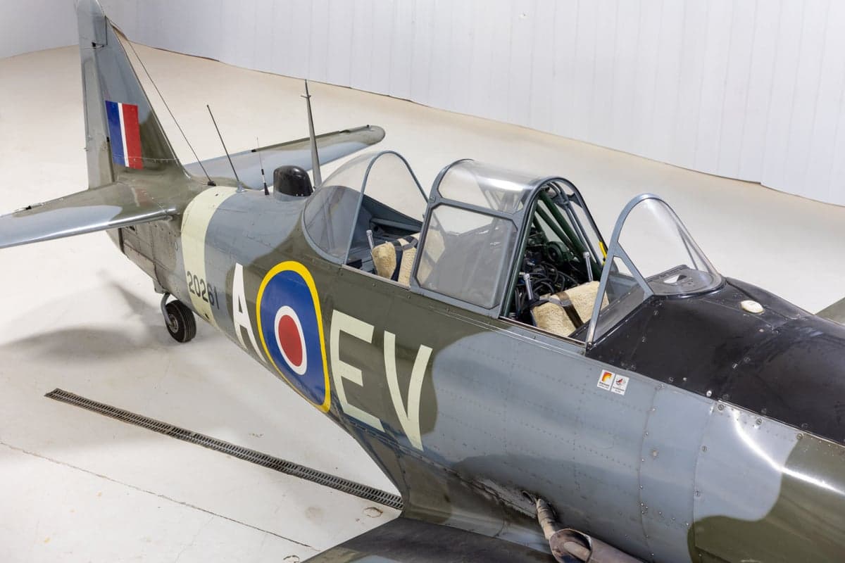 A high-angle close-up of the cockpit and mid-fuselage of a vintage military trainer aircraft with a camouflage paint scheme, parked in a hangar.
