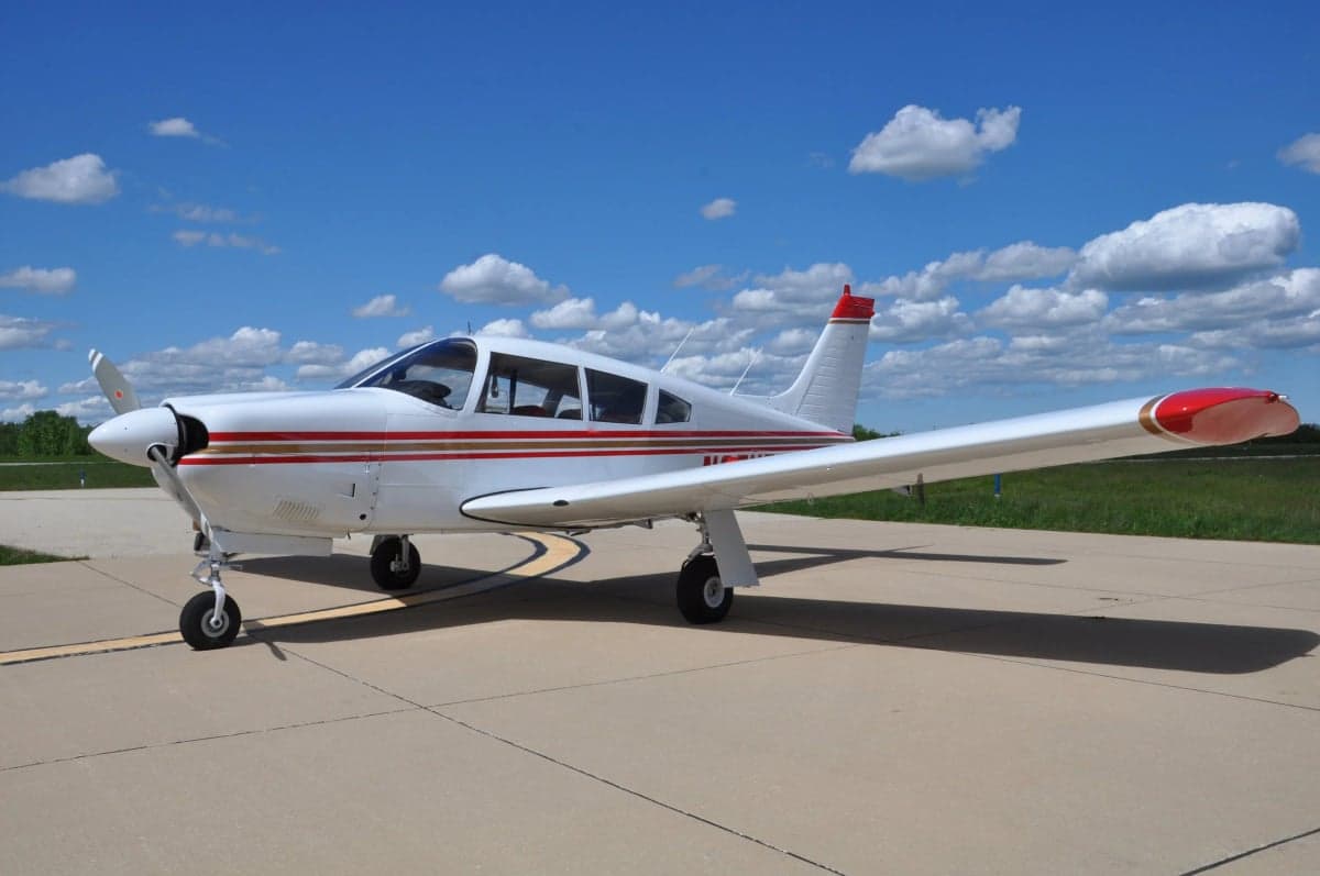 A white single-engine aircraft with red and gold stripes on a tarmac under a blue sky.