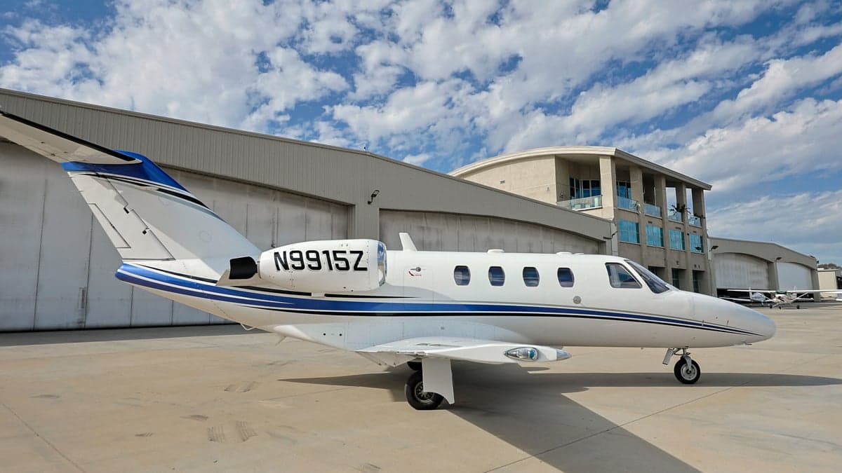 A white and blue Cessna Citation CJ1 aircraft with registration N9915Z parked on the tarmac in front of a hangar.