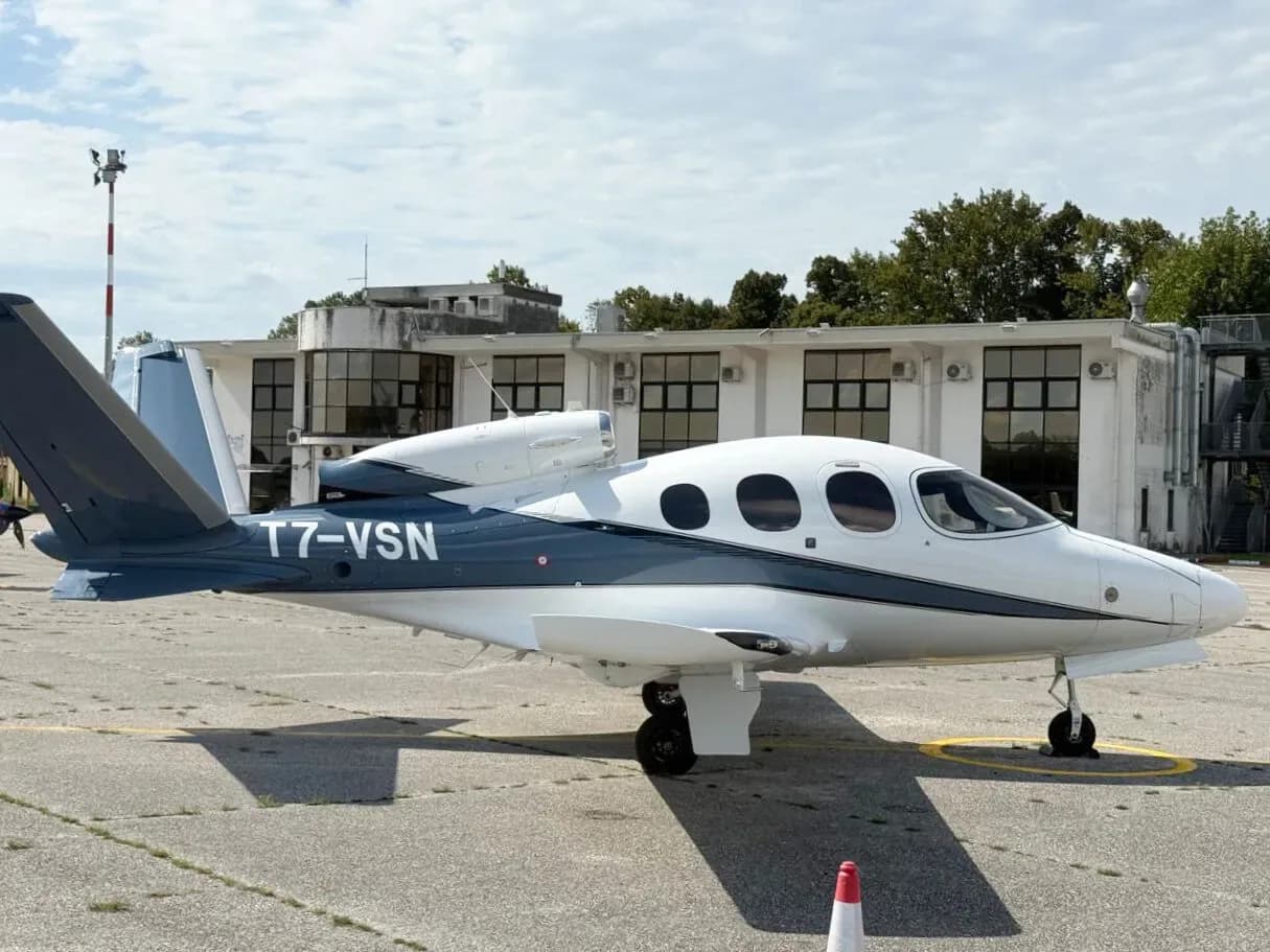 A white and dark blue Cirrus Vision Jet (T7-VSN) parked on an asphalt apron with airport buildings in the background.