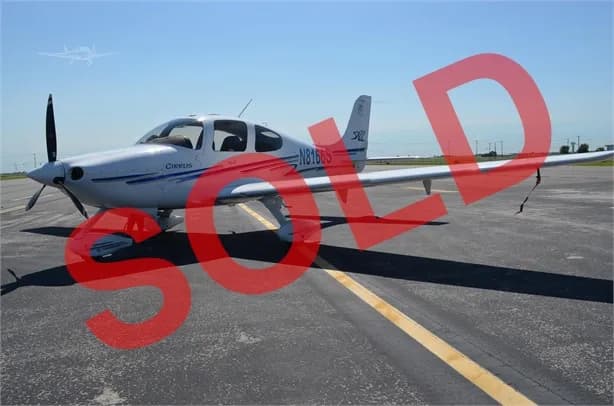 A white Cirrus SR22 aircraft with blue stripes is parked on an asphalt tarmac under a clear sky, viewed from the front-right.