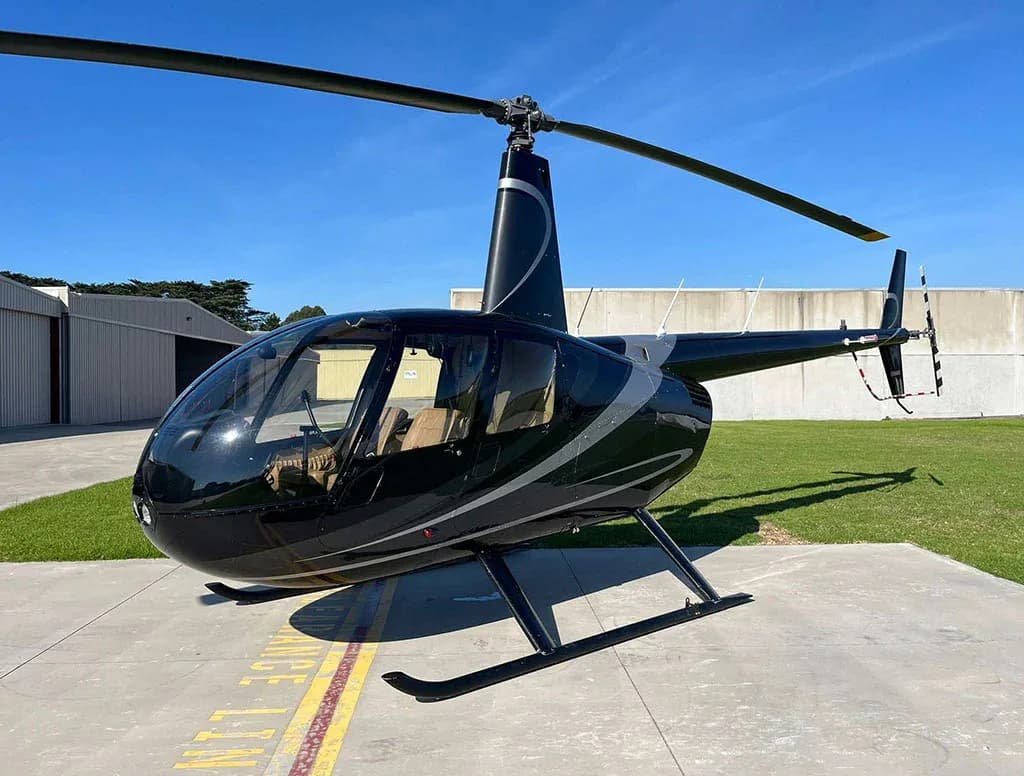 A black Robinson R44 helicopter with silver stripes on a tarmac, viewed from the front-right.