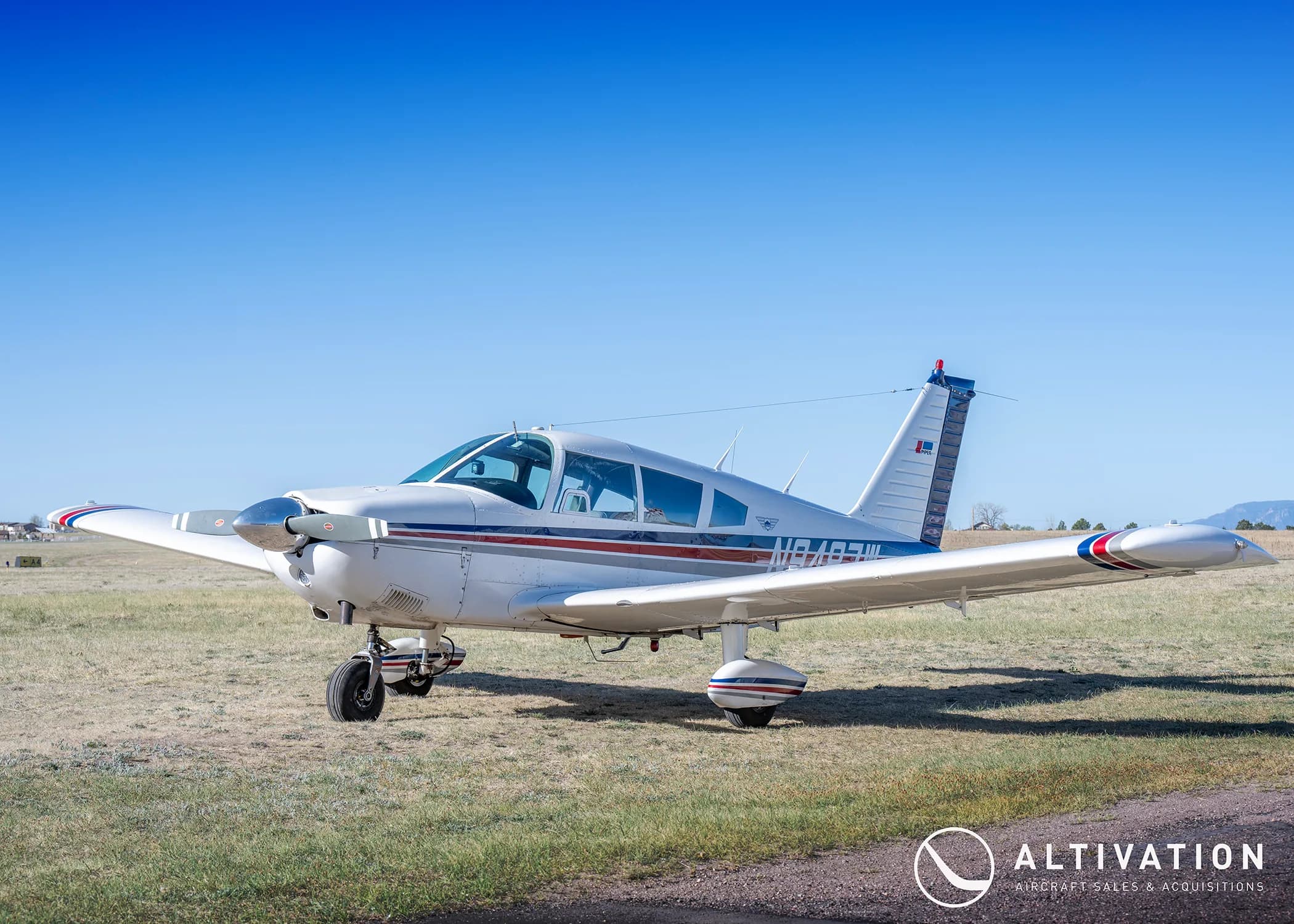 A white Piper Cherokee aircraft with red and blue stripes on a grassy field under a clear blue sky.