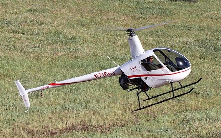 A white Robinson R22 helicopter flying over a grassy field.