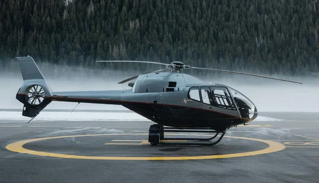 A sleek grey and black helicopter with a red stripe sits on a helipad, with a misty, snow-covered forest in the background.