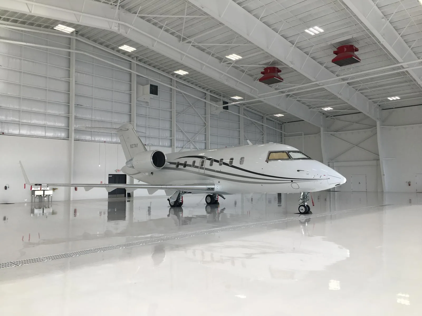 A white business jet with black and grey stripes, tail number N27AY, is parked in a brightly lit hangar.