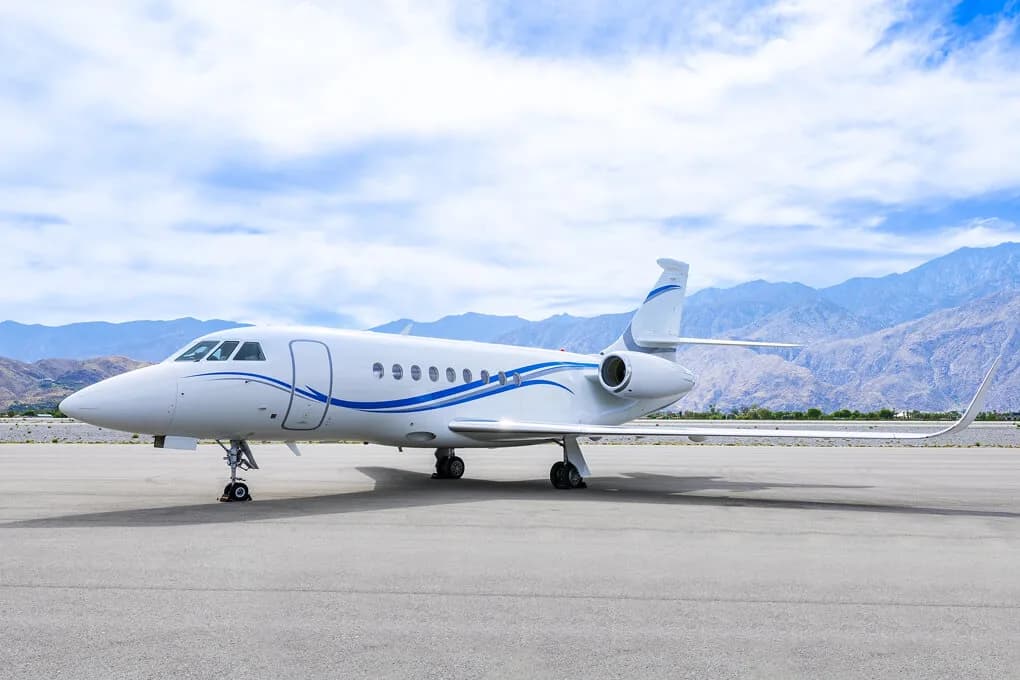 A white Dassault Falcon 2000LX business jet with blue stripes on the fuselage is parked on an airfield with mountains in the background.
