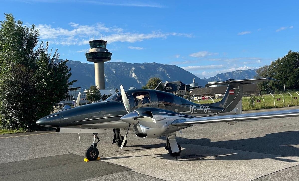 Diamond DA40 aircraft with registration OE-FGE parked on the tarmac with an airport control tower in the background.