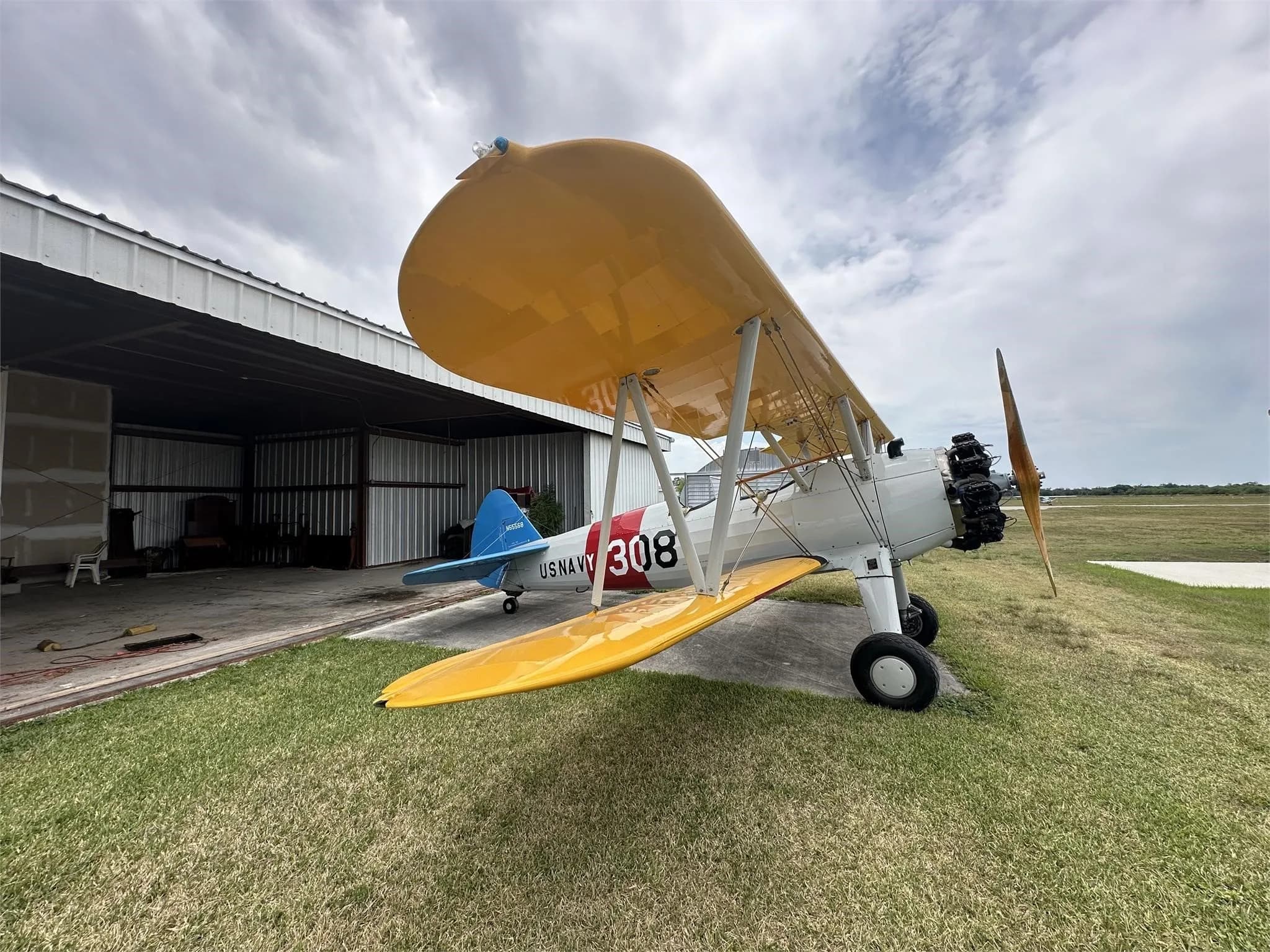 A white biplane with yellow wings and "US NAVY 308" markings is parked on grass next to an open hangar under a cloudy sky.
