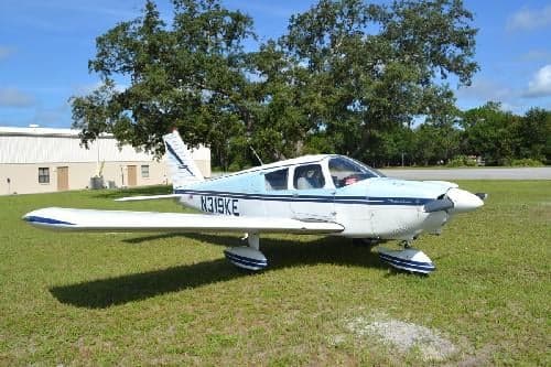 A white and blue Piper PA-28-140 Cherokee sits on a grassy field.
