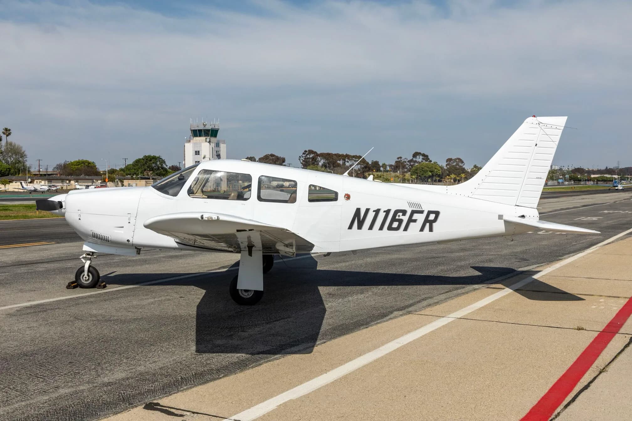 A white Piper PA-28-181 Archer III aircraft, registration N116FR, parked on an airfield with a control tower in the background.