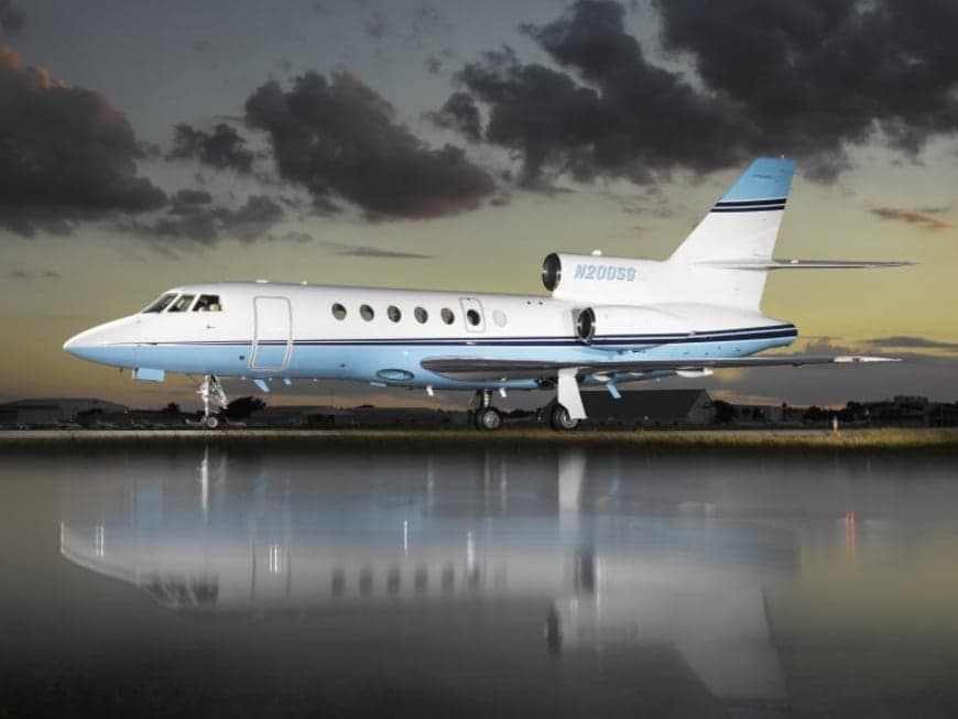 A Falcon jet with blue and white paint scheme is reflected in the water on the tarmac at sunset.