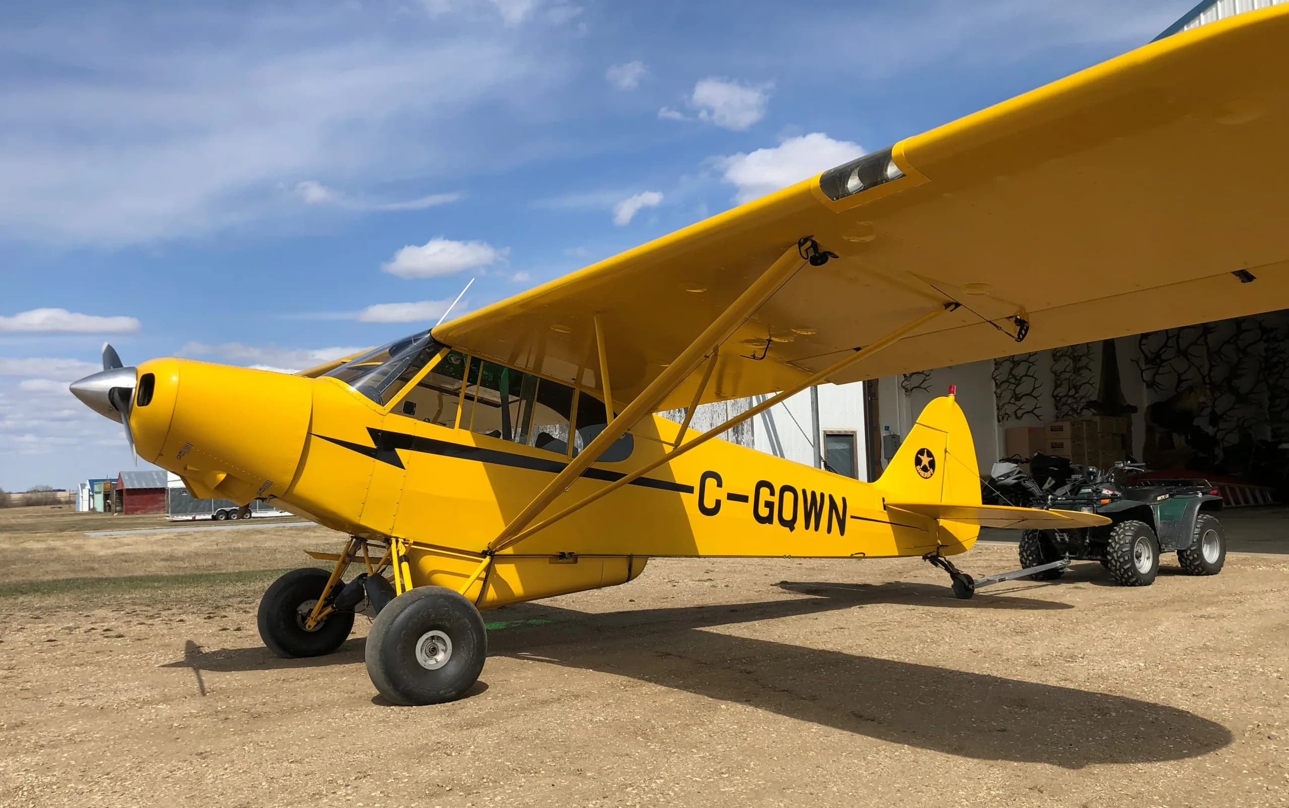 A bright yellow Piper Cub-style aircraft with large tundra tires, registration C-GQWN, is parked on a dirt field next to a hangar with an ATV.