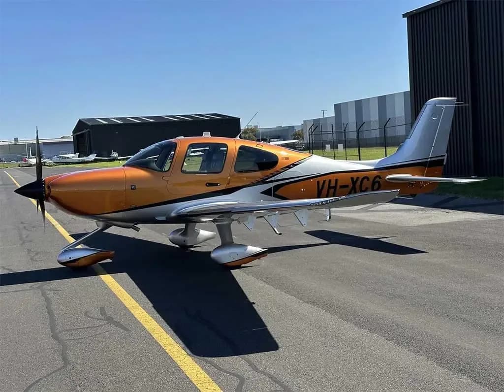 A side view of an orange and silver Cirrus SR22 aircraft with registration VH-XC6 parked on an airport tarmac.