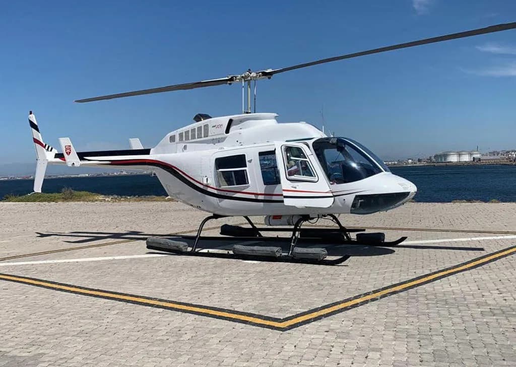 A white helicopter with black and red stripes on a helipad by the ocean under a clear blue sky.