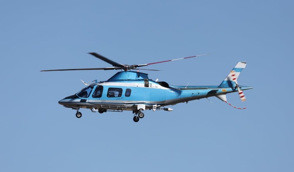 A blue and white AgustaWestland AW109 helicopter in flight against a clear blue sky.