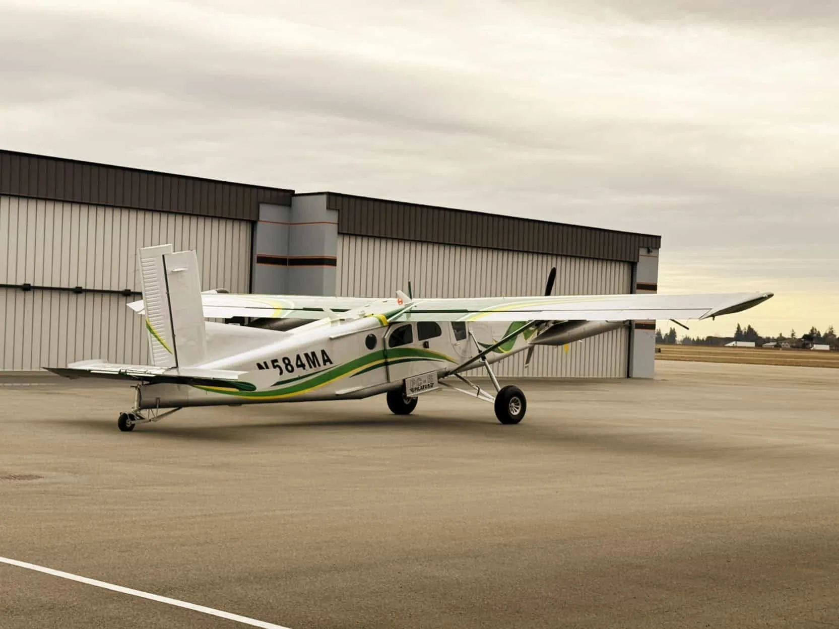 A Pilatus PC-6 Turbo Porter aircraft with a white and green paint scheme is parked on the tarmac in front of a hangar.