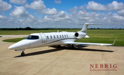 A white business jet with a black stripe, registration N30AB, parked on a tarmac with green fields and a partly cloudy sky in the background.
