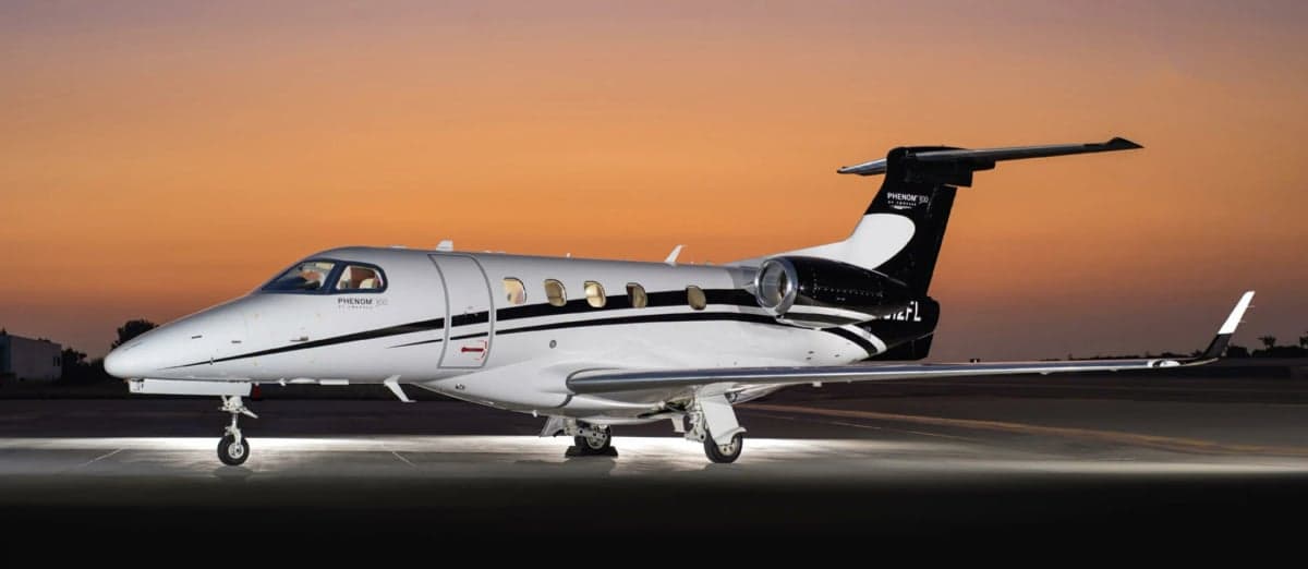 A Phenom 300 aircraft with a white and black paint scheme is parked on the tarmac at sunset.