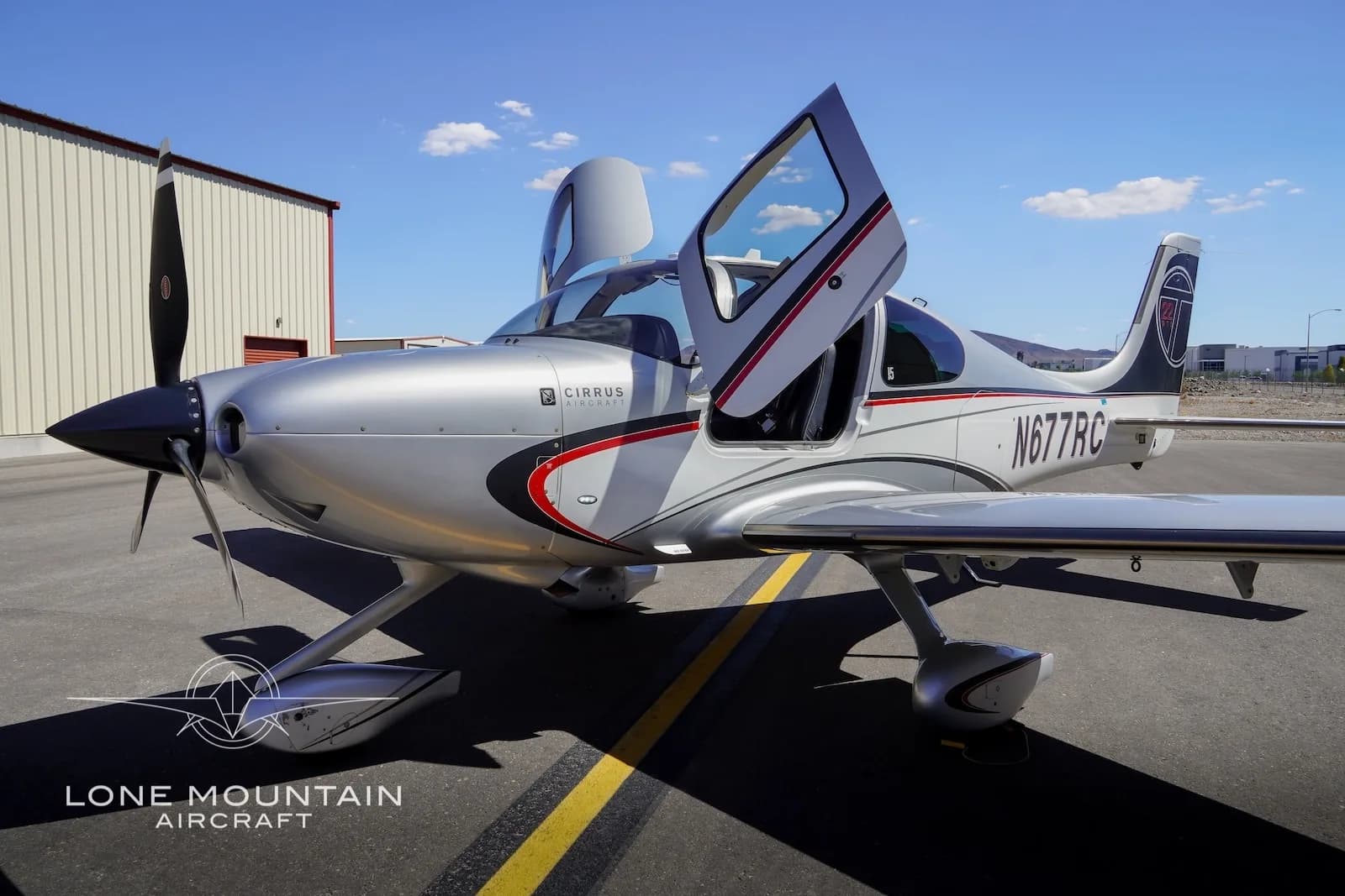 A silver Cirrus SR22 aircraft with red and black accents, registration N677RC, parked on an airfield with its gull-wing doors open.