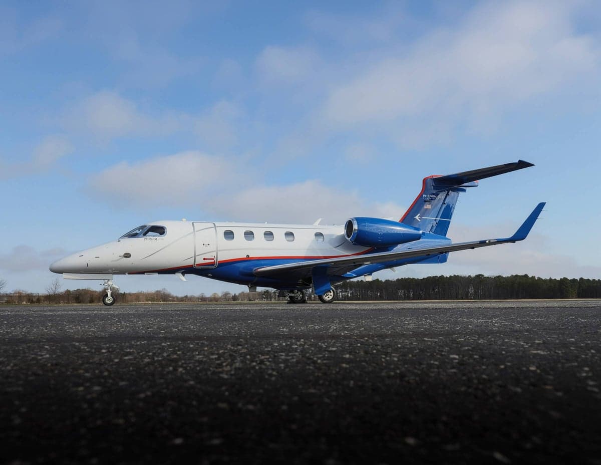 A Phenom 300E aircraft is parked on the tarmac with a blue and white paint scheme.