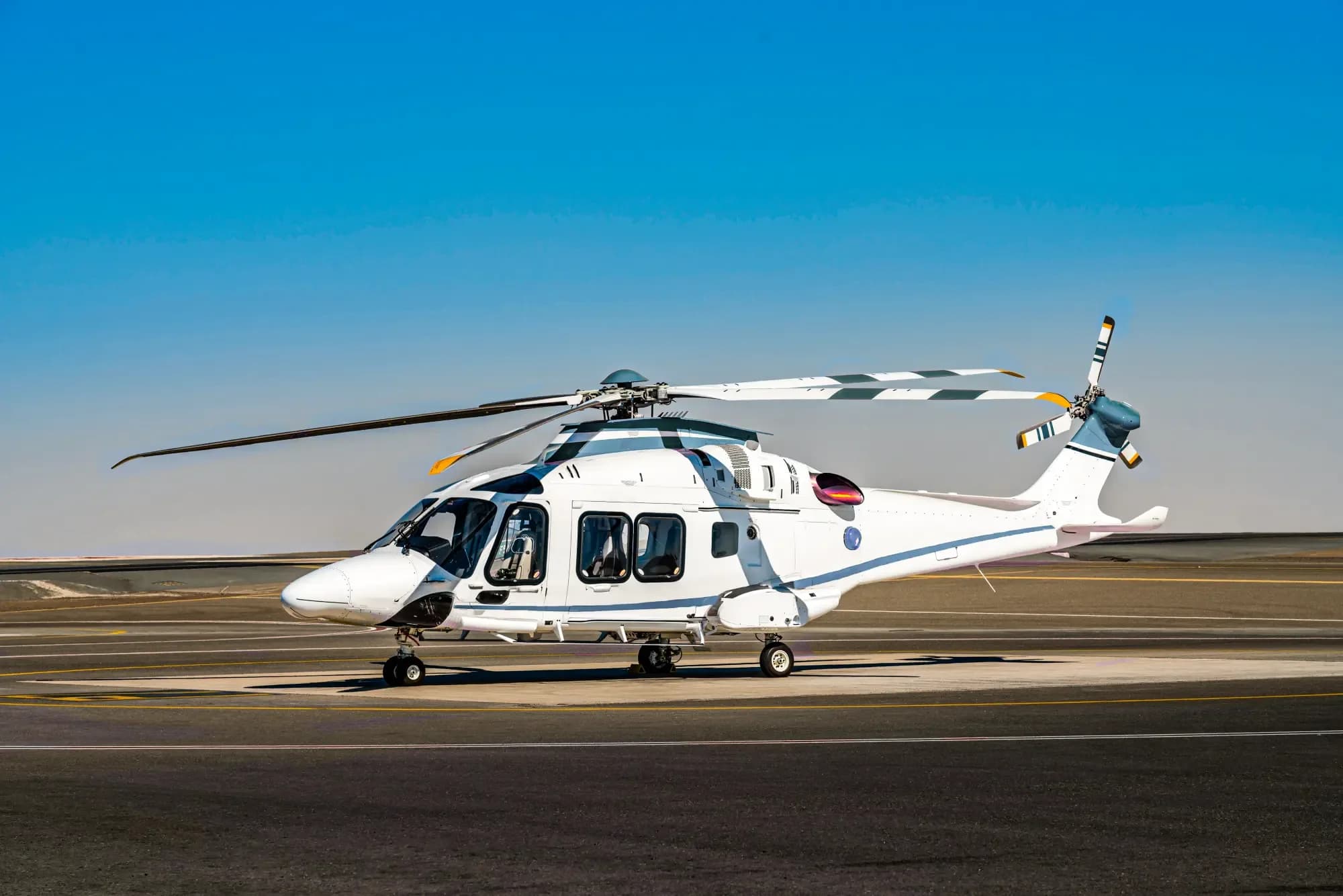 A white helicopter with blue and grey accents sits on an airport tarmac under a clear blue sky.
